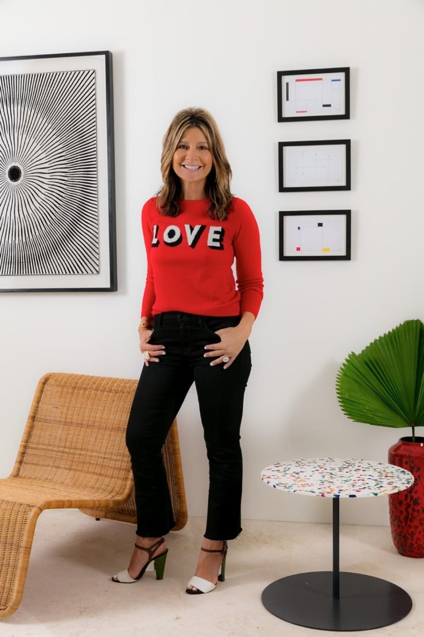 Person in a red sweater with "LOVE" text, standing by a wicker chair and a small table, with modern art in the background.