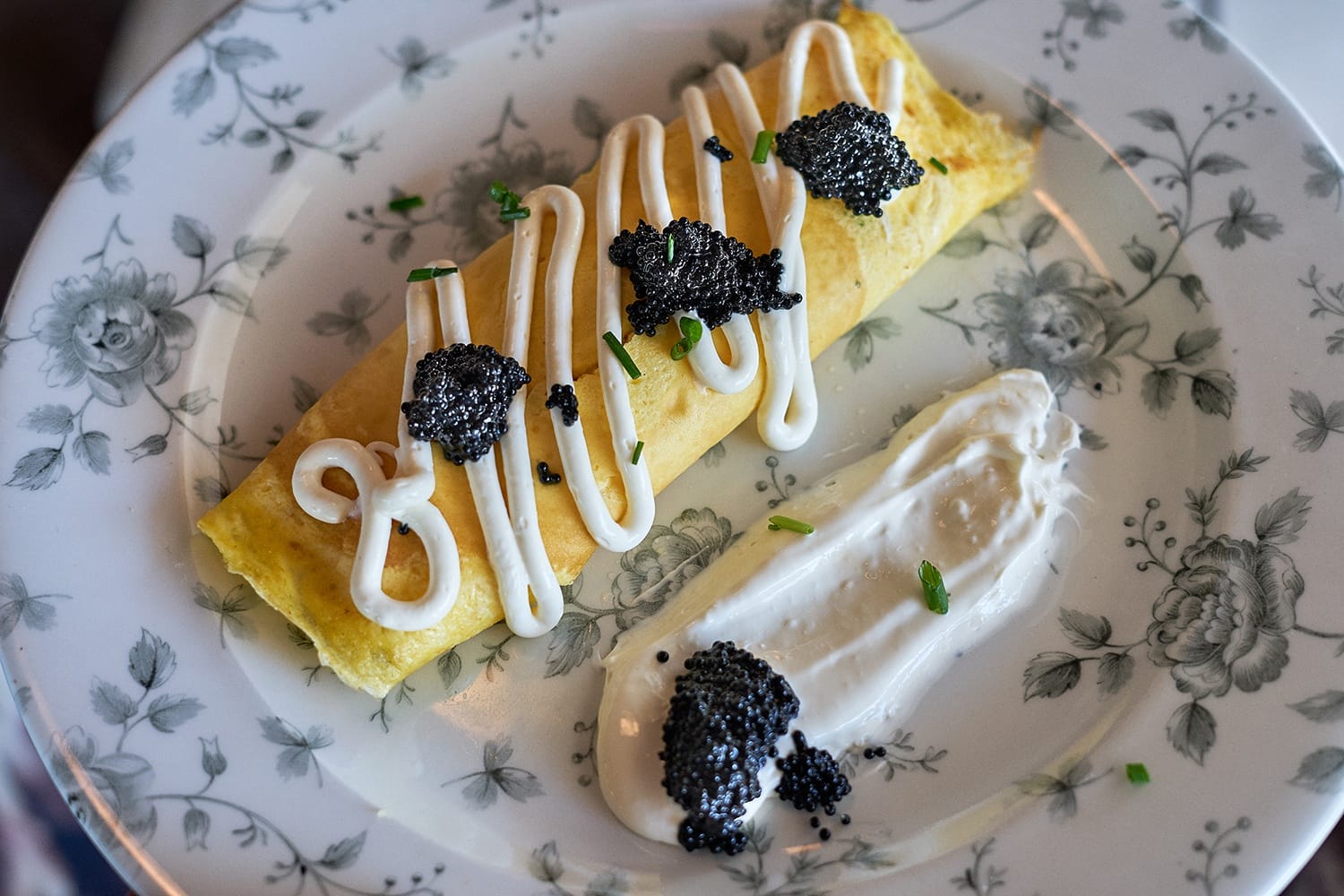 Plate with a rolled crepe topped with white sauce and caviar, alongside a dollop of sour cream, garnished with chives.