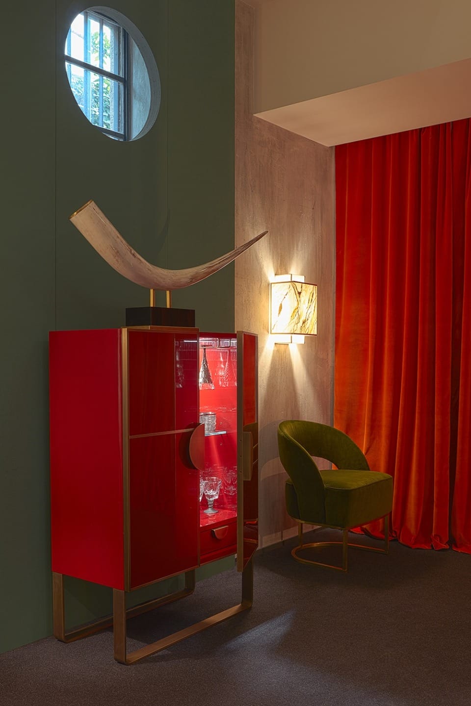 Stylish interior with red cabinet, glassware, green chair, curved art piece, round window, orange curtains, and wall light.