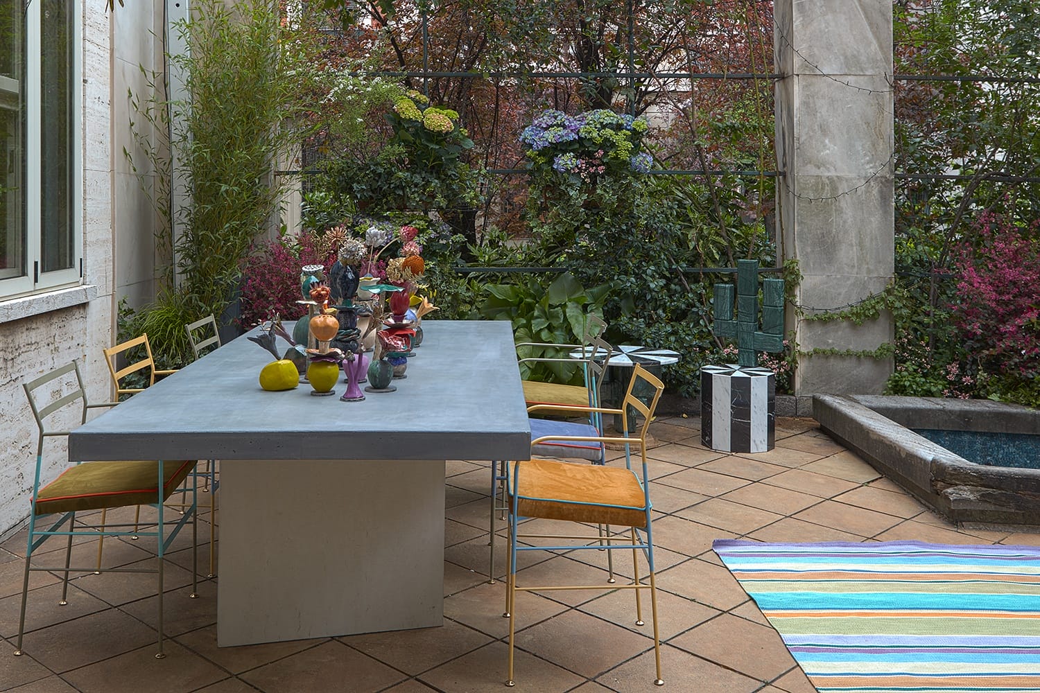 Outdoor patio with a modern table, colorful chairs, plants, and a small fountain set against a background of lush greenery.
