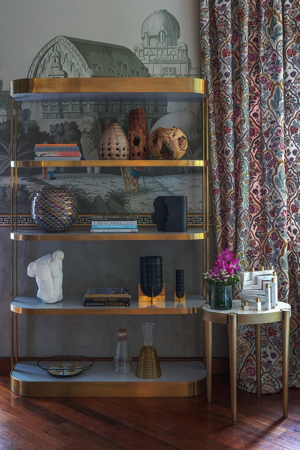 Elegant shelving with books, artistic sculptures, vases, and decorative items beside a table with flowers, in a richly patterned room.