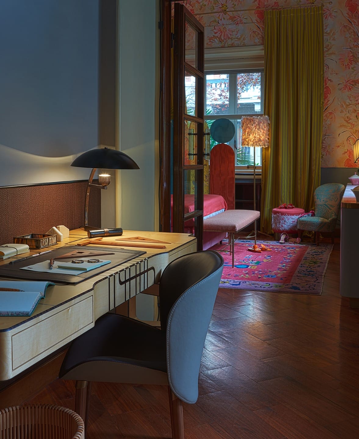 Vintage-inspired study with wooden desk, modern chair, desk lamp, and a cozy living area with colorful decor in the background.