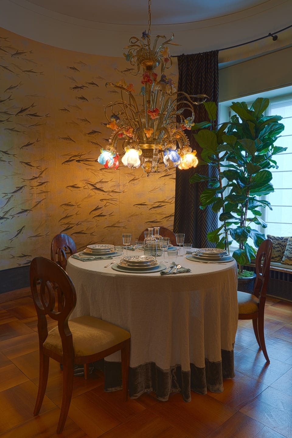 Elegant dining room with round table set for four under a colorful chandelier, surrounded by decorative wallpaper and plants.