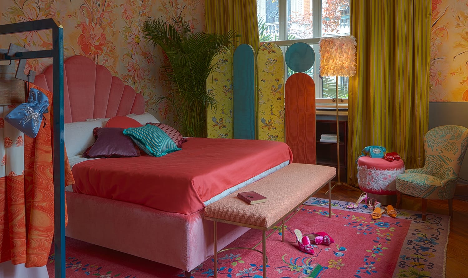 Colorful bedroom with pink bed, floral wallpaper, vibrant decor, and various textiles creating a cozy, eclectic atmosphere.