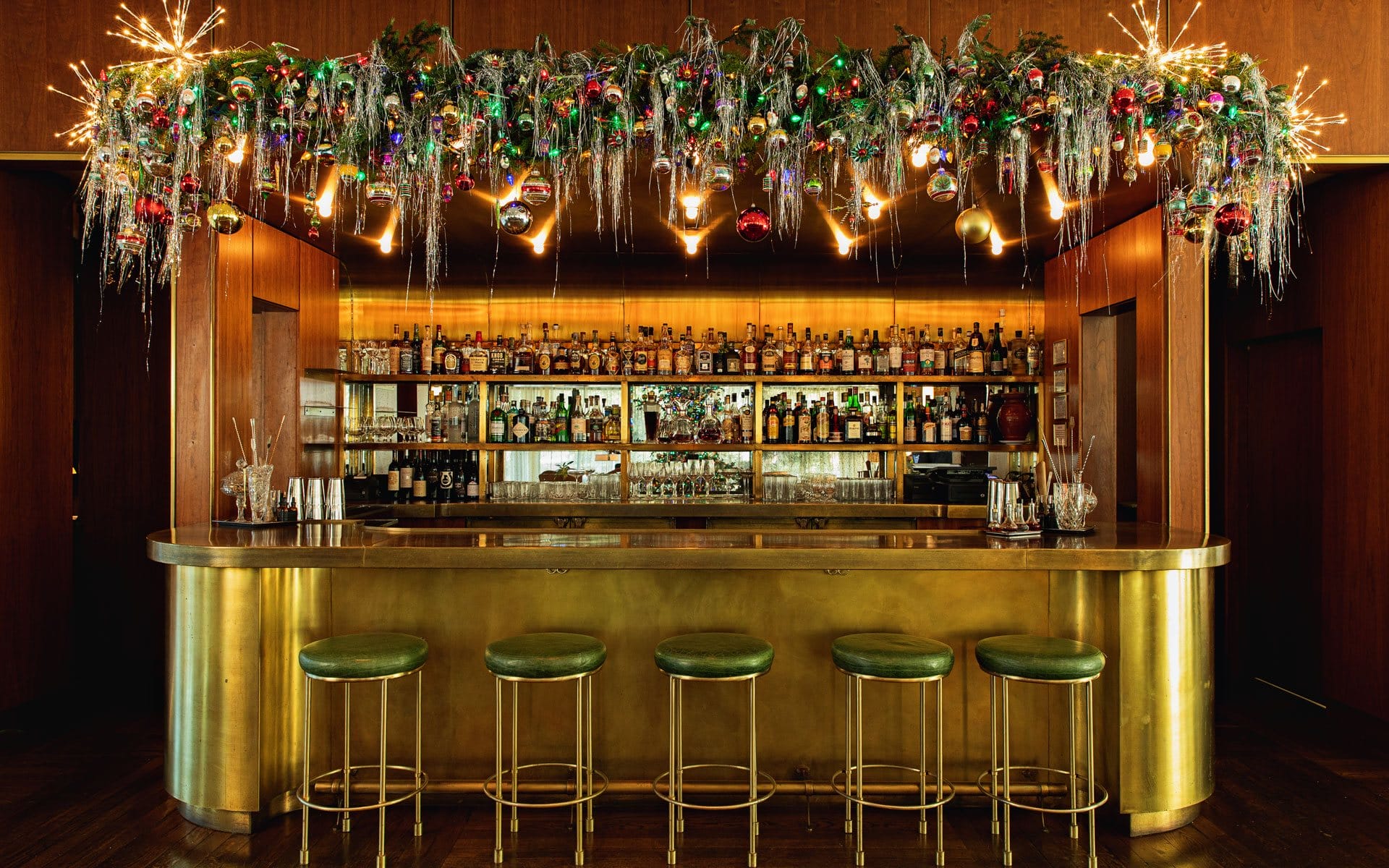 Festive bar decorated with greenery and ornaments above a sleek counter with green stools, surrounded by a warm, wooden interior.