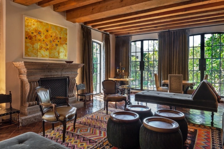 Rustic living room with wooden beams, large windows, fireplace, colorful rug, and eclectic furniture arrangement.