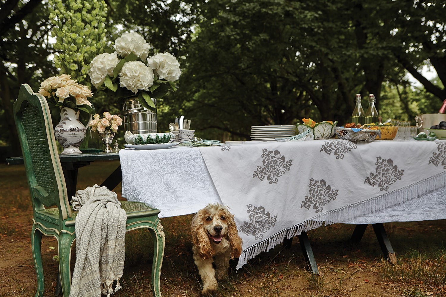 Outdoor table setting with flowers, white tablecloth, dishes, and a playful dog under the table in a wooded area.