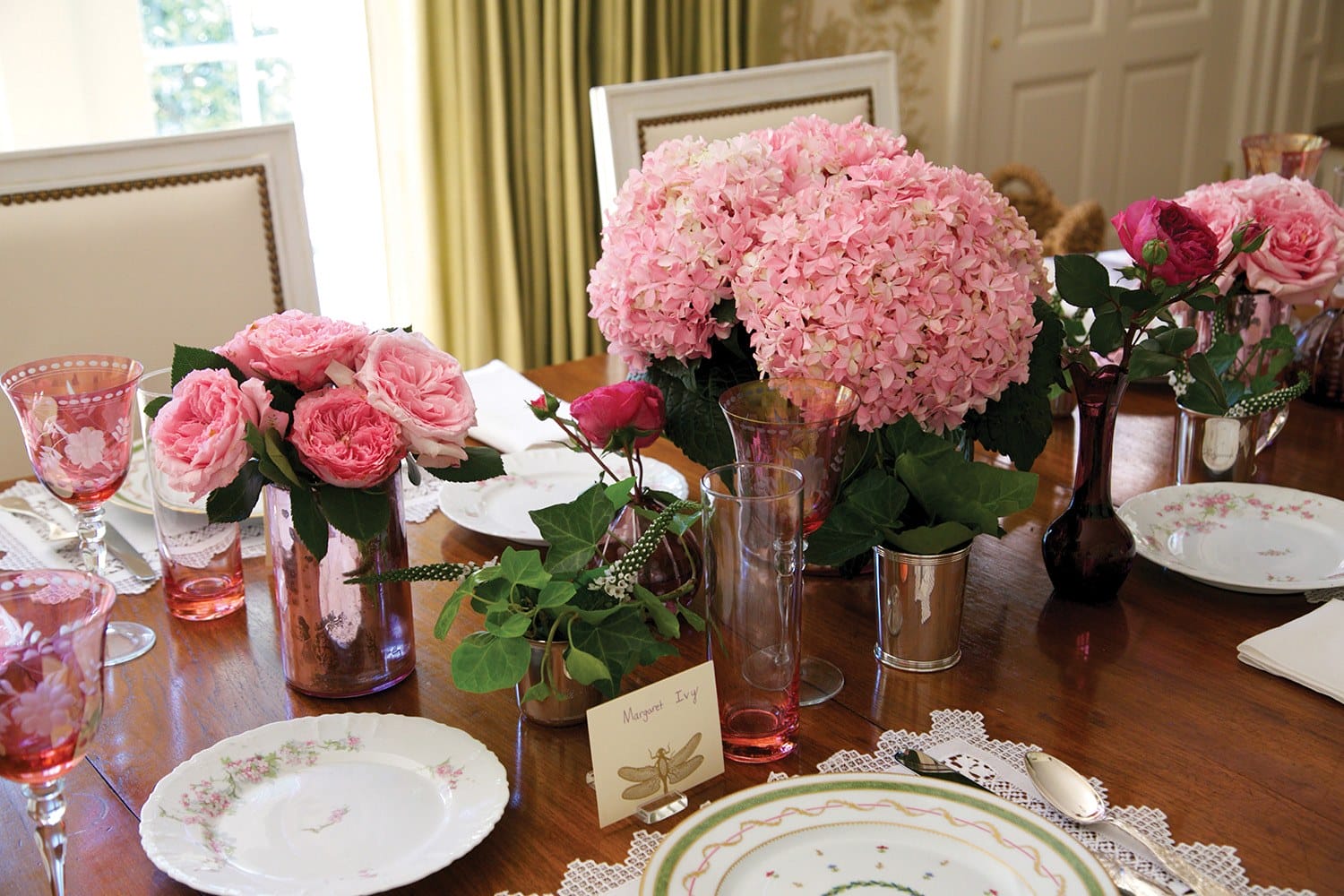 Elegant dining table set with pink hydrangeas and roses, fine china, and crystal glasses in a warmly lit room.