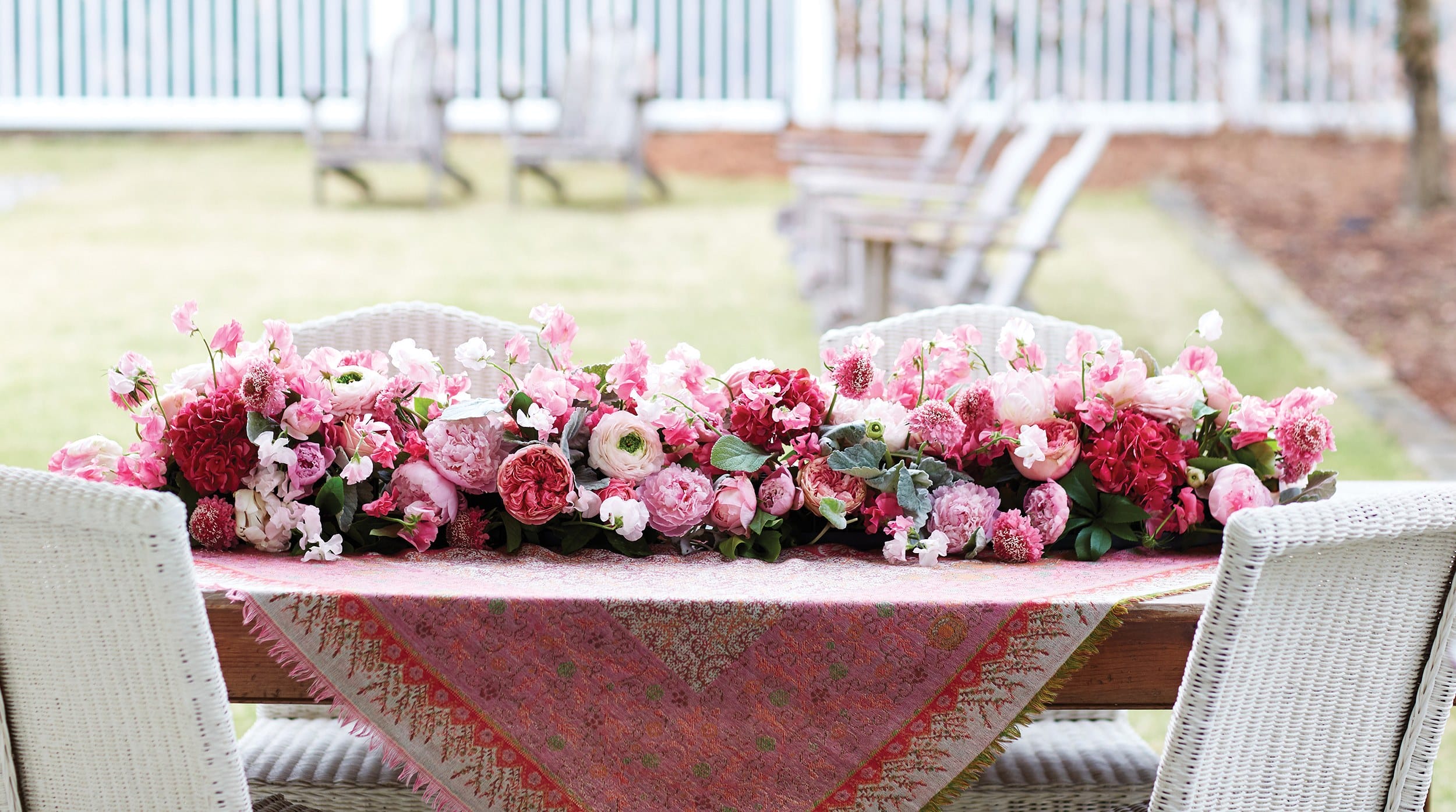 Floral centerpiece on an outdoor table with pink and red peonies, surrounded by chairs and a patterned tablecloth.