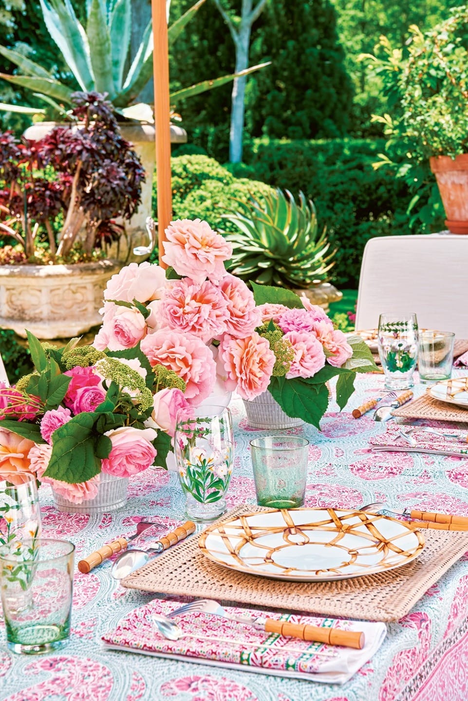 Outdoor dining table set with floral centerpiece, patterned tablecloth, and plates, surrounded by lush garden greenery.