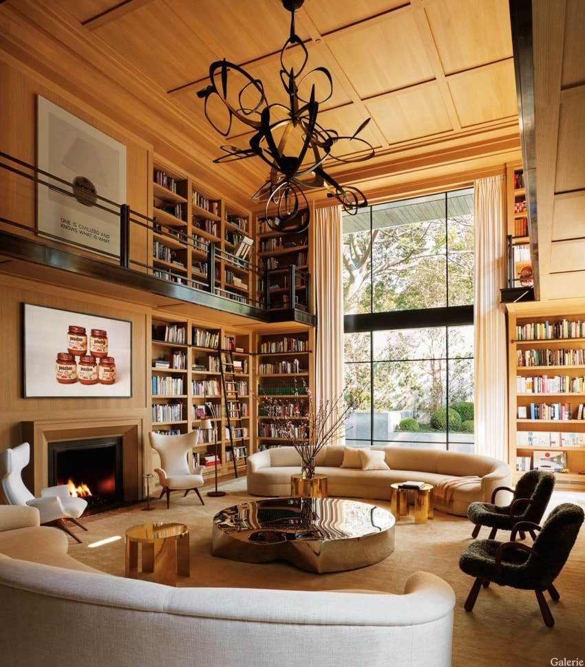 Luxurious living room with large windows, bookshelves, modern chandelier, soft seating, and a fireplace.