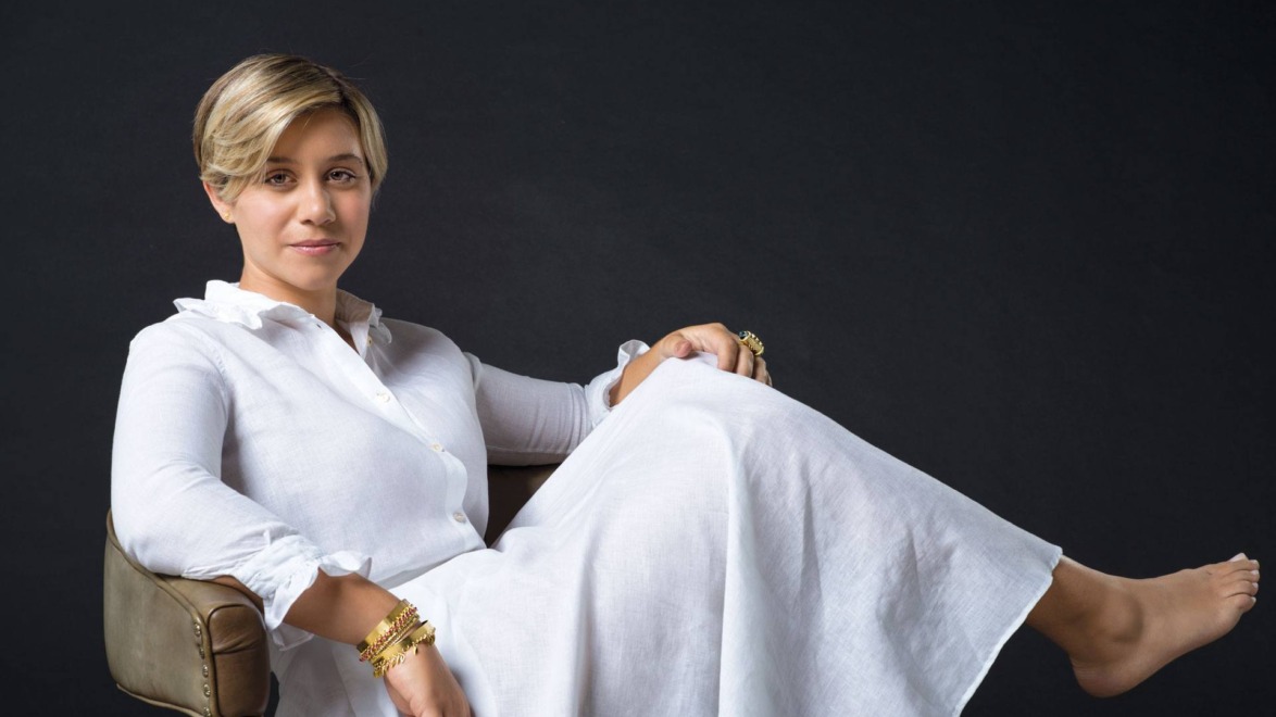 Person sitting in a chair wearing a long white dress against a dark background.
