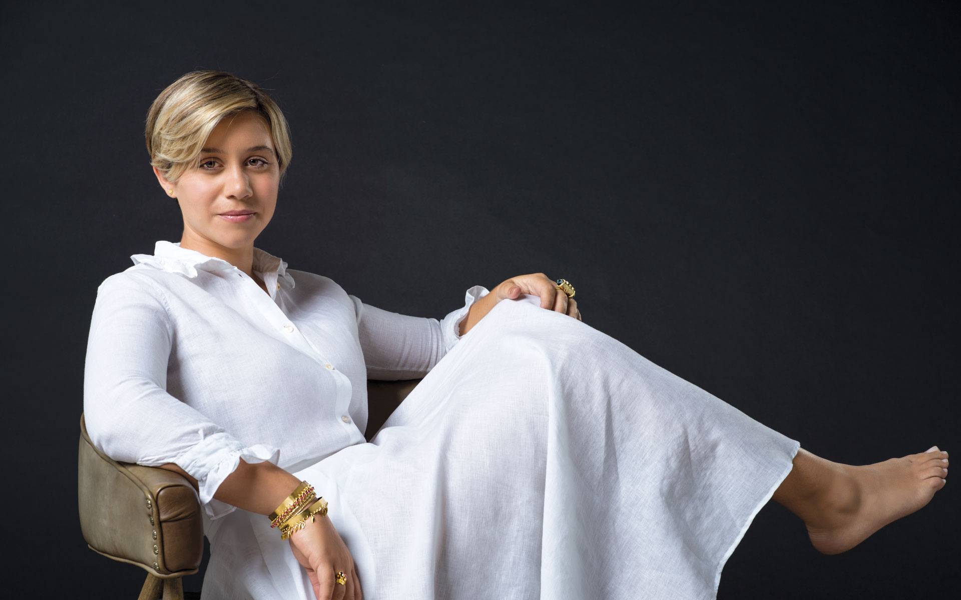 Person sitting in a chair wearing a long white dress against a dark background.