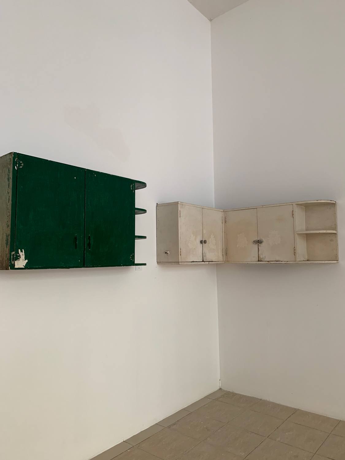 Two vintage wall-mounted cabinets, one green and one beige, in a minimalistic room with white walls and tiled flooring.