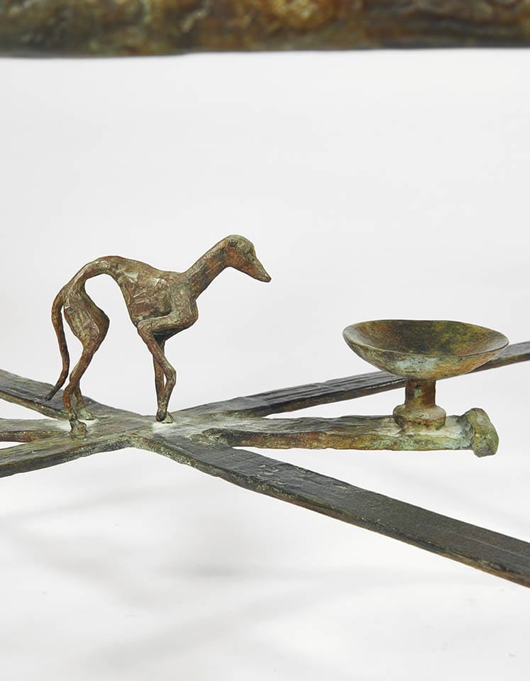 Sculpture of a thin horse beside a small round dish on a cross-shaped base.