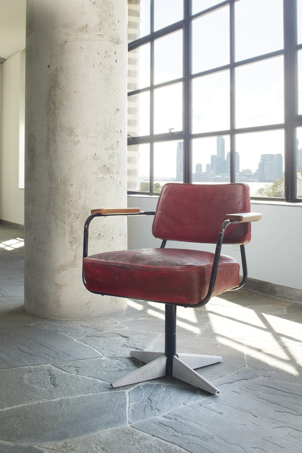 Red vintage chair with metal frame placed in a modern industrial-style room with large windows and cityscape view.