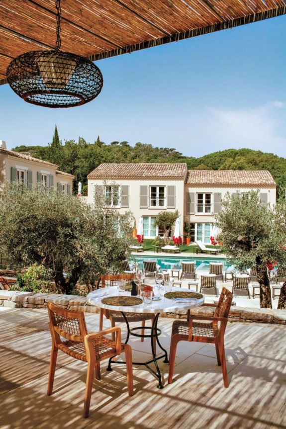 Outdoor patio with table and chairs overlooking a pool and villa surrounded by trees under a clear blue sky