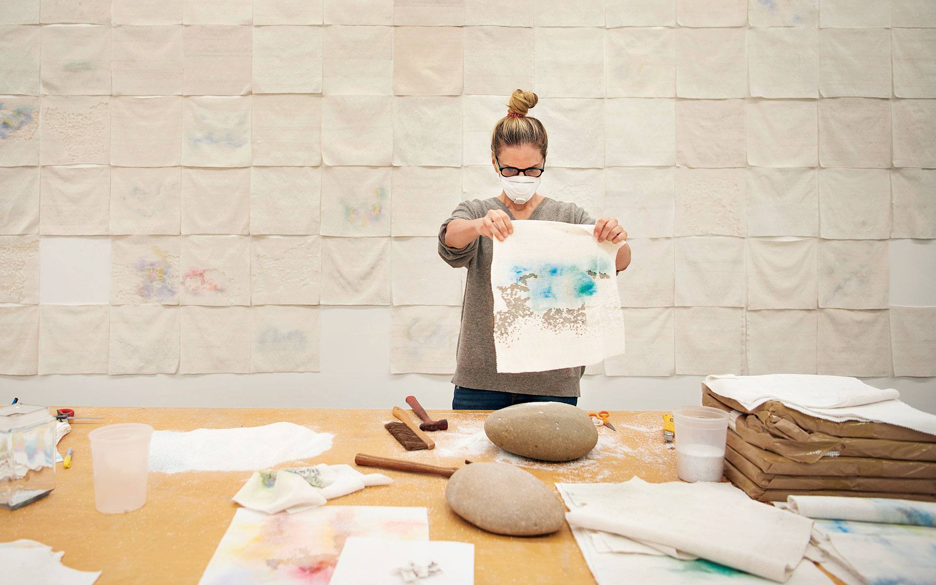 Artist wearing a face mask examining a canvas in a studio with materials on the table and art pieces on the wall.