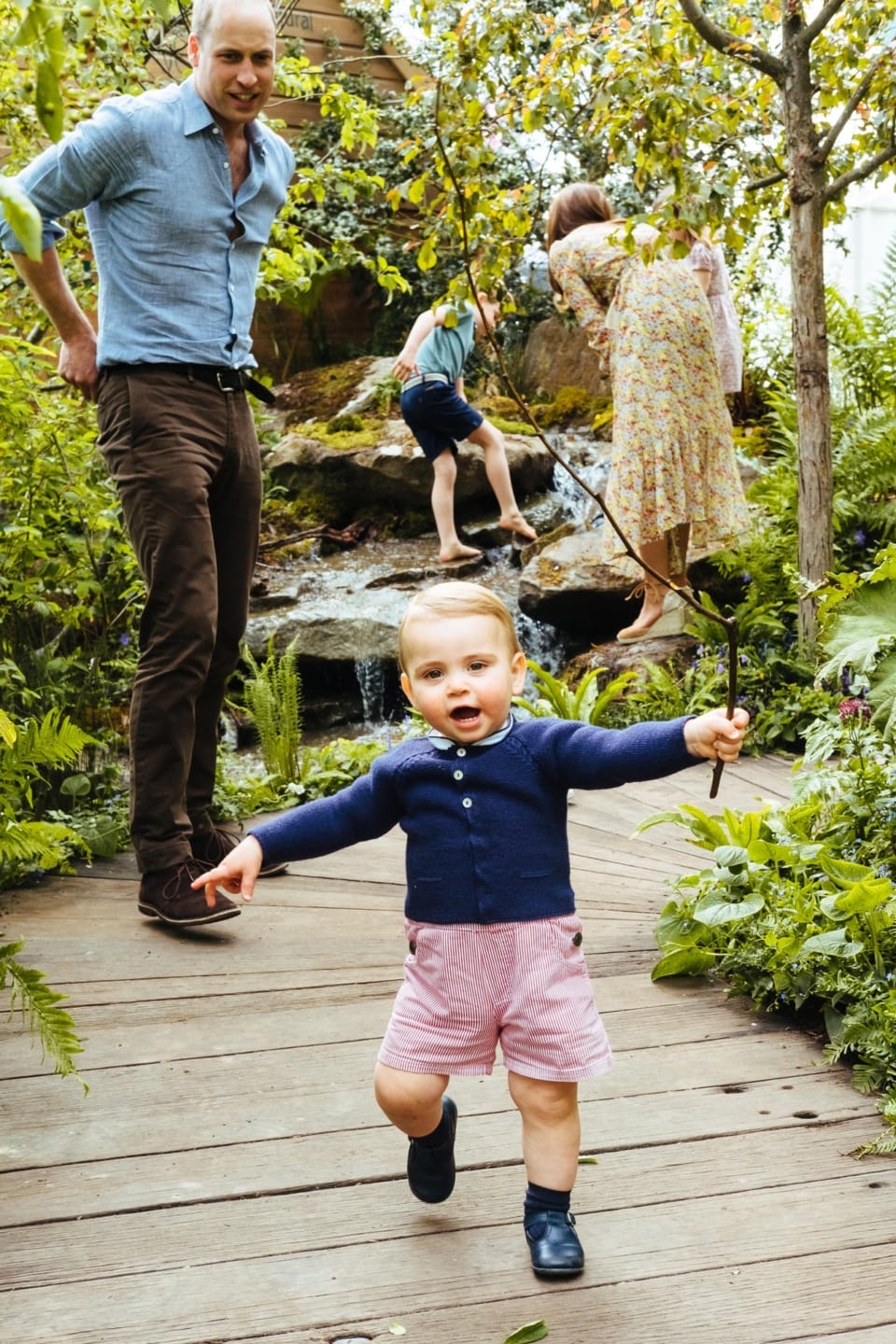 Toddler walking on a wooden path with adults and children in a lush garden setting.