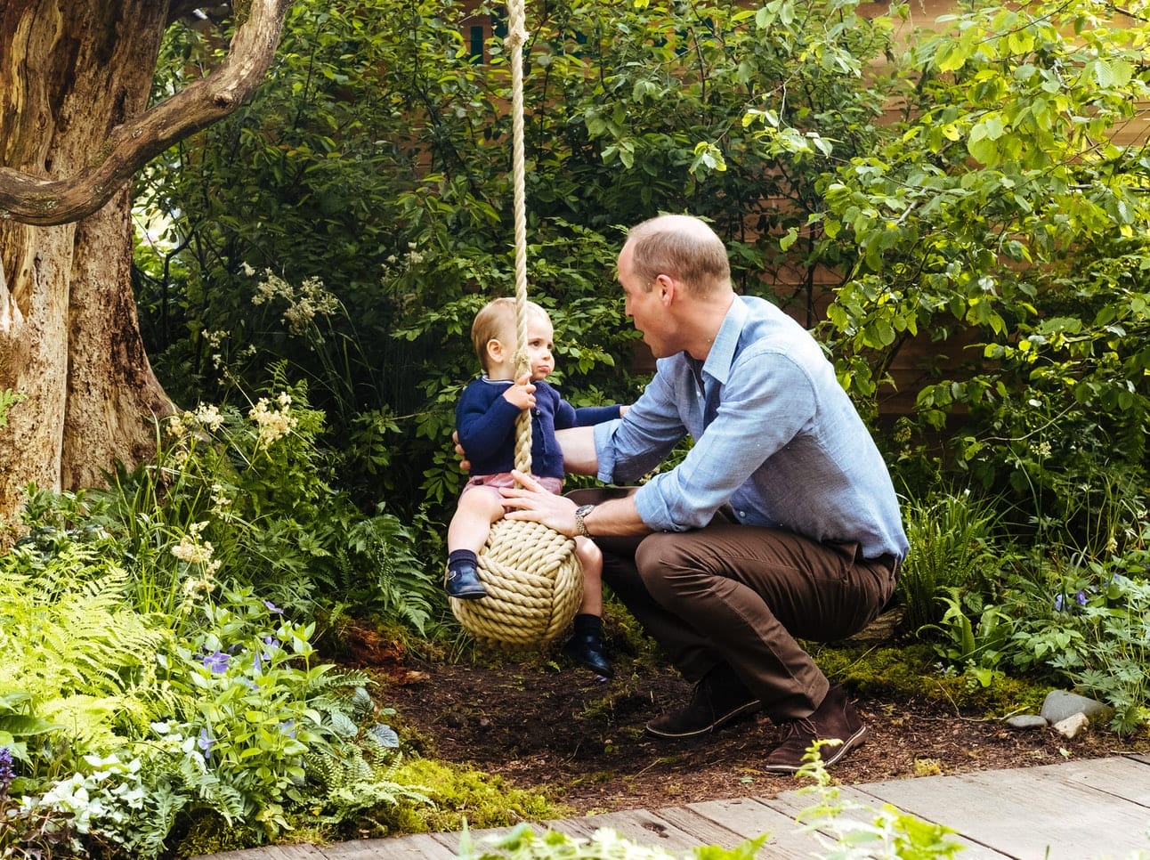 Man kneeling and holding a rope swing with a small child seated on it, surrounded by lush green garden foliage.