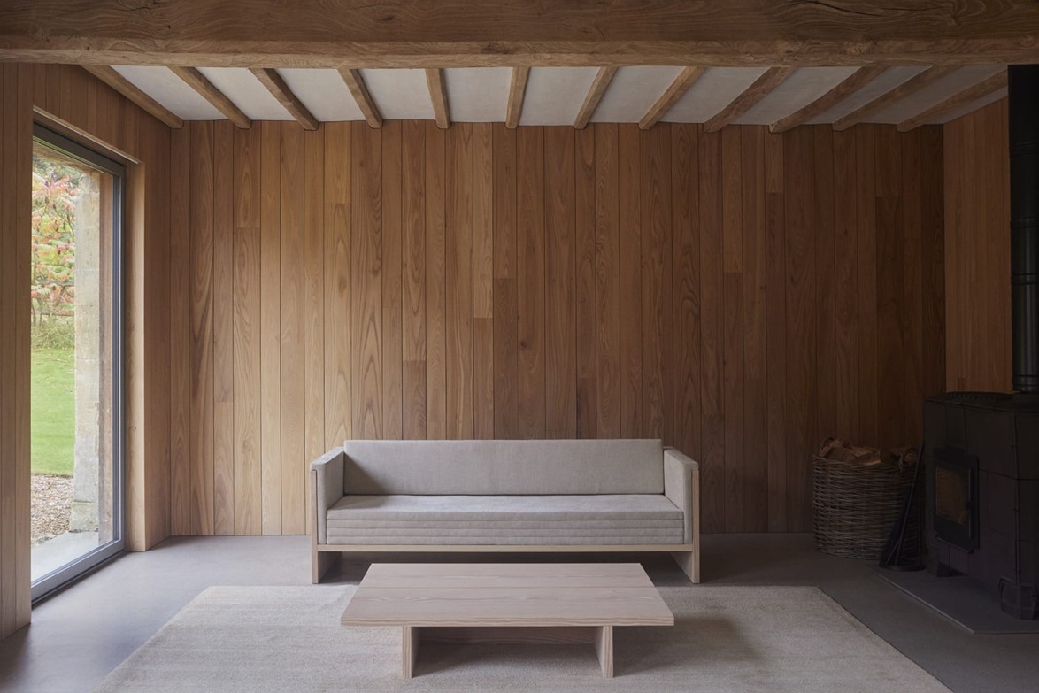 Minimalist living room with wooden walls, a beige sofa, a simple coffee table, a large window, and a wood-burning stove.