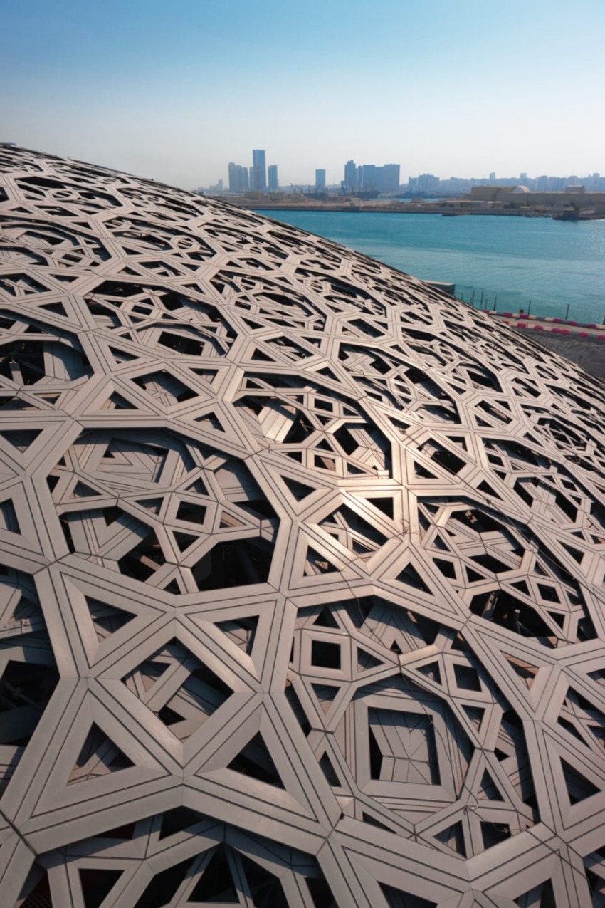 Geometric patterned exterior of a modern building with a city skyline and waterfront in the background, under a clear blue sky.