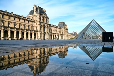 Louvre Museum courtyard with glass pyramid and building reflection in water puddle under clear blue sky.
