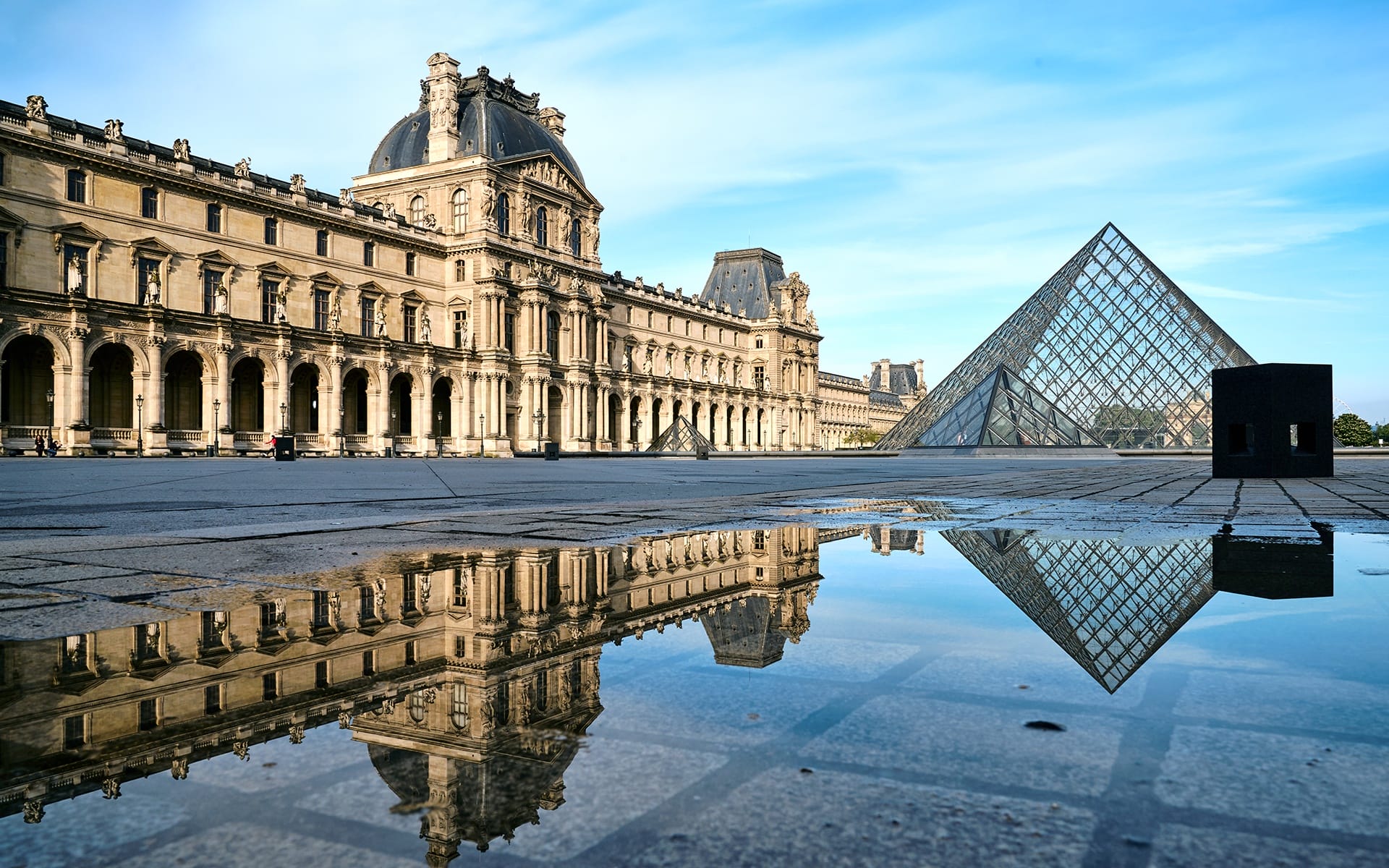Louvre Museum courtyard with glass pyramid and building reflection in water puddle under clear blue sky.