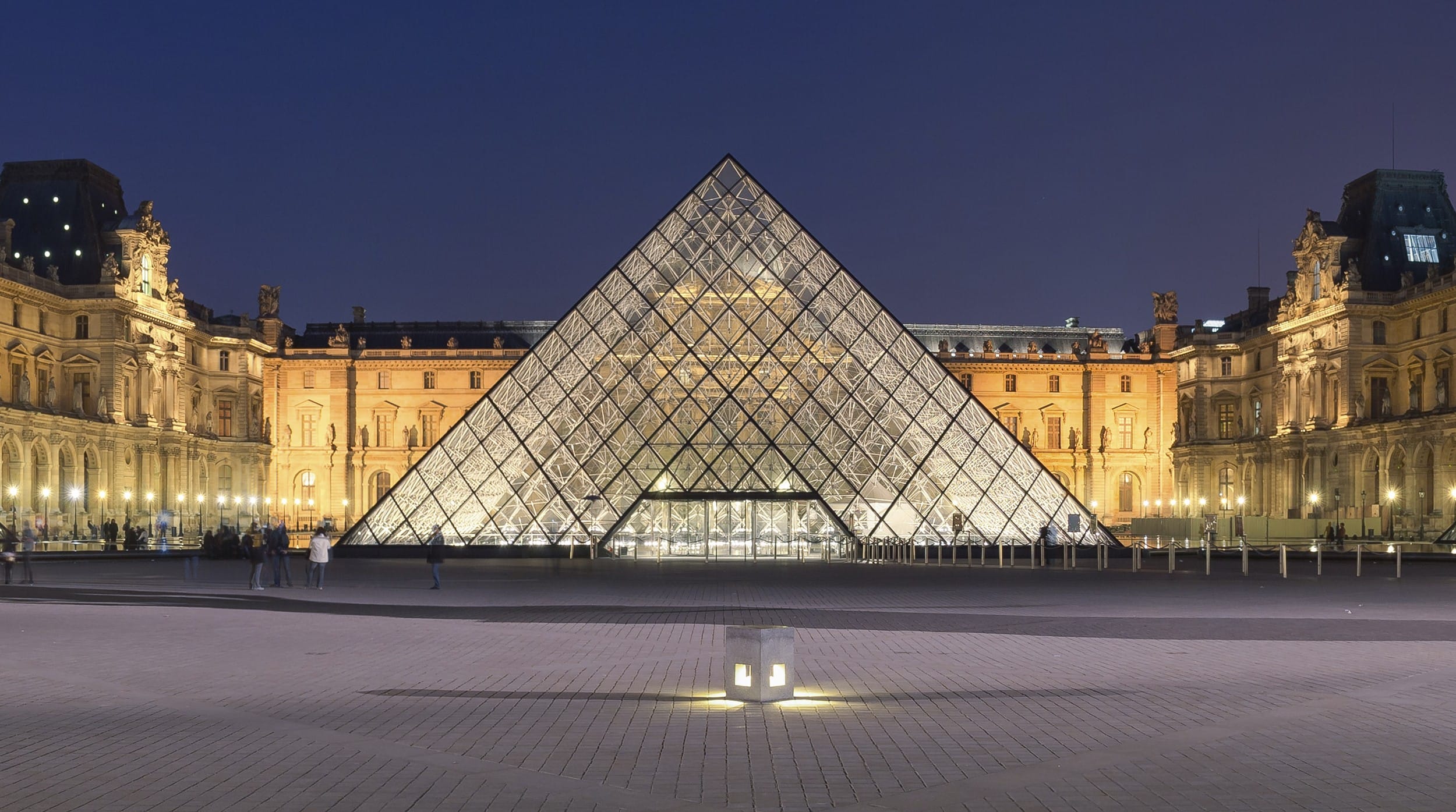 Louvre Pyramid illuminated at night in front of the historic Louvre Museum in Paris with a clear evening sky.