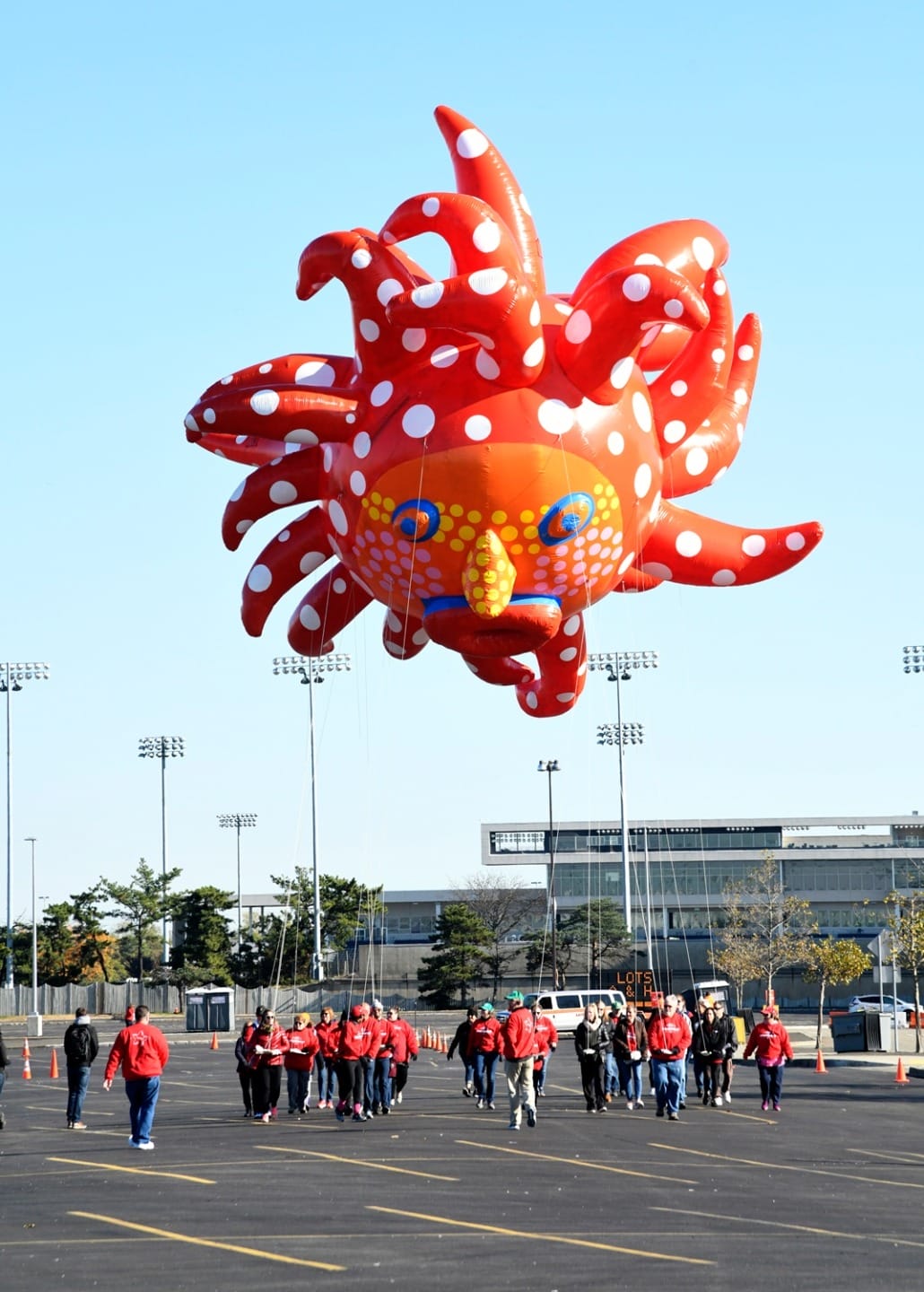 Large red octopus balloon with white spots floats above a group of people walking in a parking lot.