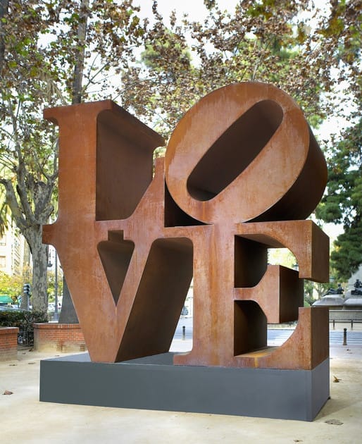 Large rust-colored "LOVE" sculpture in a park setting with trees in the background.