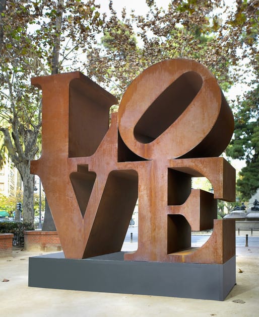 Large rust-colored "LOVE" sculpture in a park setting with trees in the background.