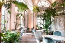 Elegant cafe interior with tall plants, arched windows, and marble tables, creating a lush and inviting atmosphere.