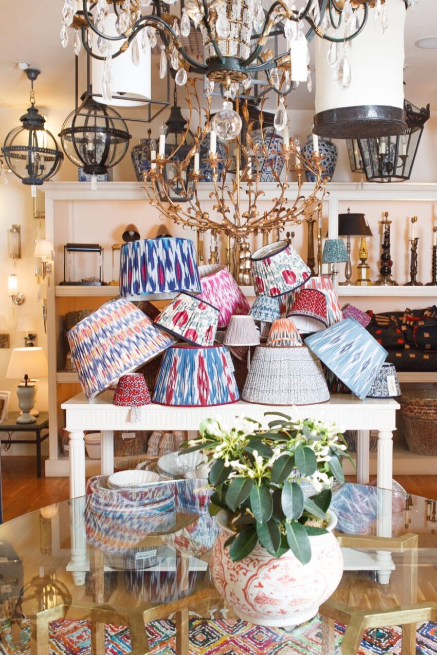 Display of colorful patterned lampshades on a white table in a brightly lit room with chandeliers and decorative lighting fixtures.