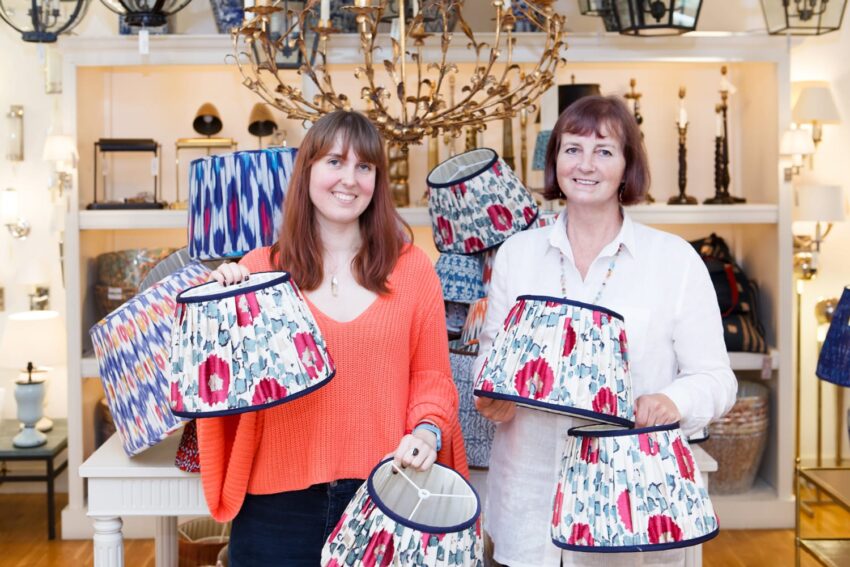 Two women smiling while holding colorful lampshades in a decorative lighting store.