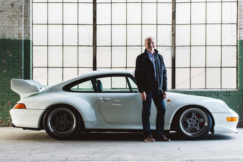 Man standing beside a classic white sports car in an industrial setting with large windows in the background.