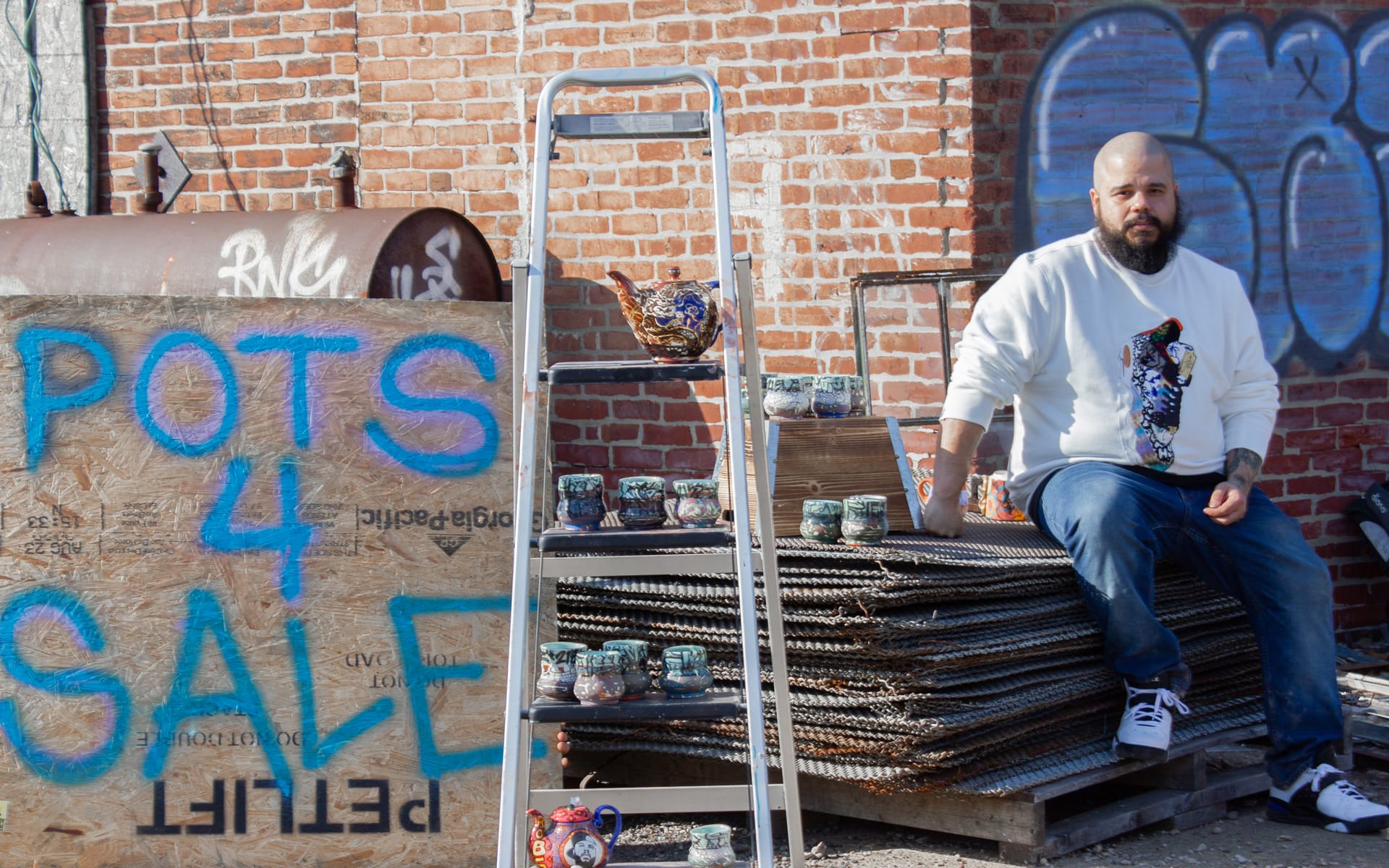 Man sitting beside a stack of metal, next to painted sign reading "Pots 4 Sale" with various ceramic pots displayed.