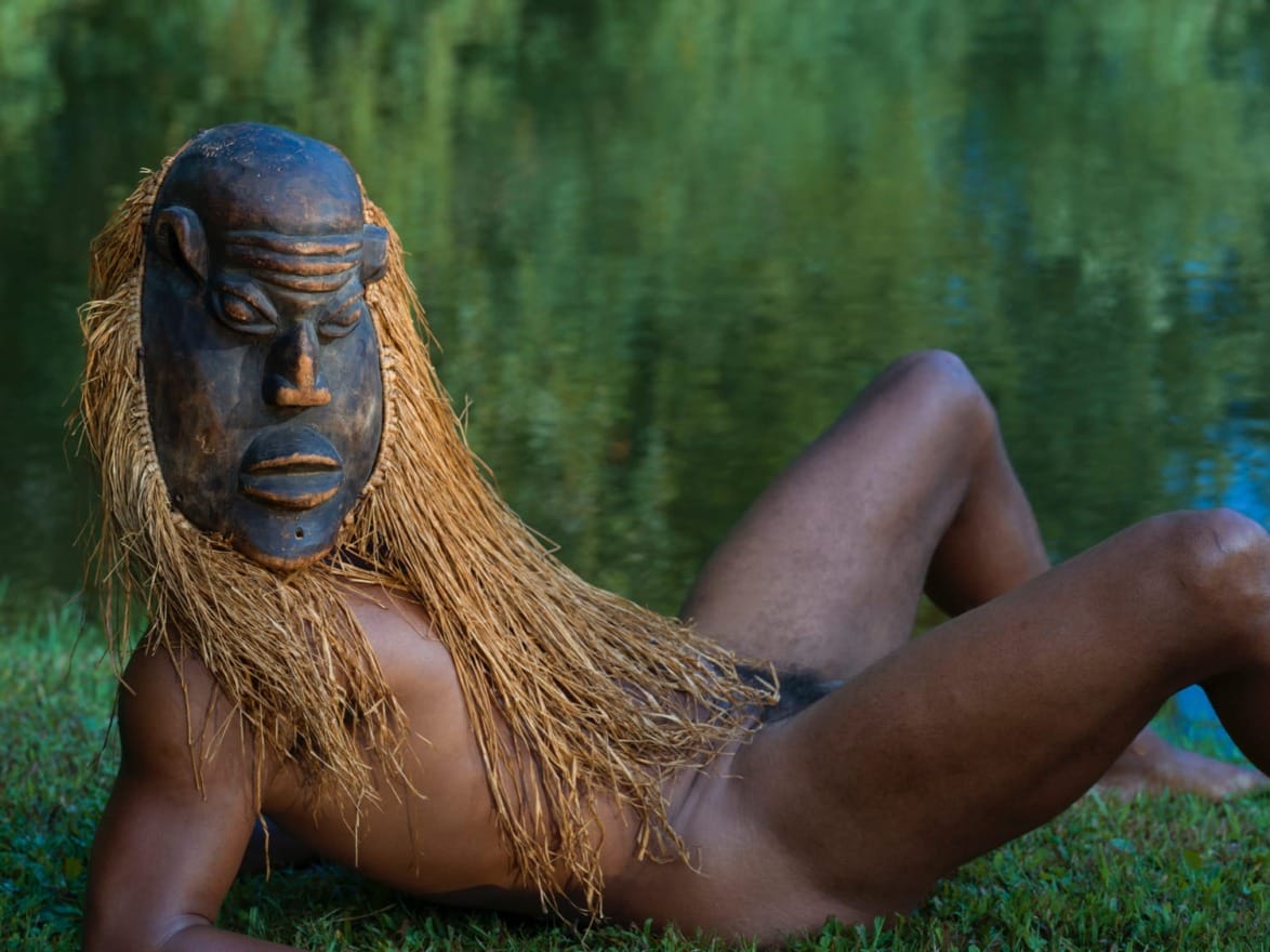 Person wearing a large mask and natural fiber costume, reclining on grass near a lake with a lush green background.