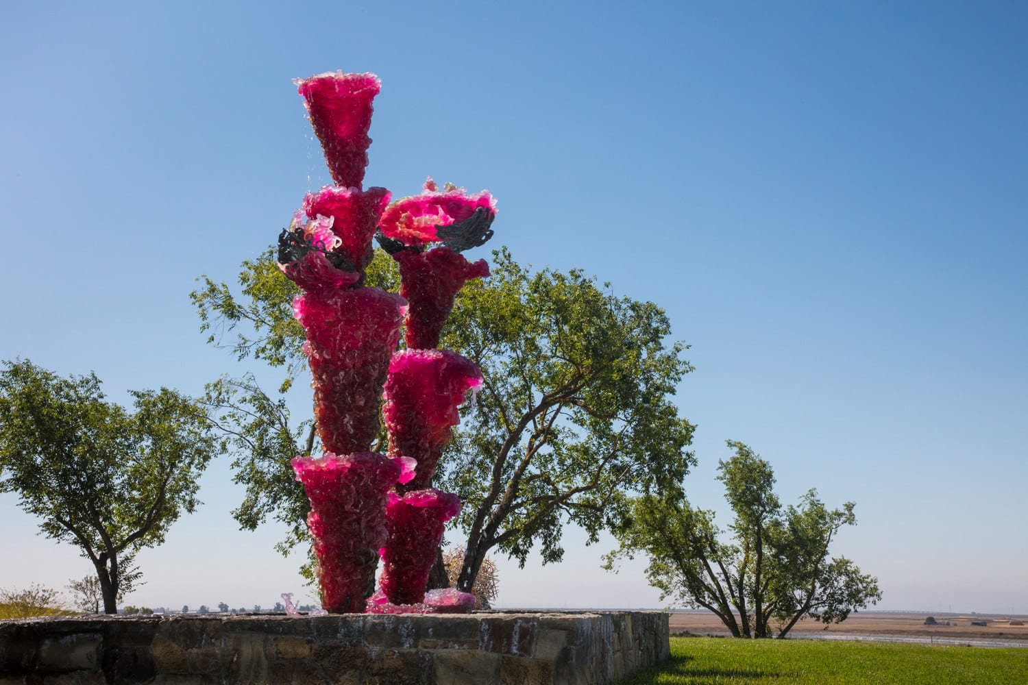 Pink glass sculpture in sunny outdoor park surrounded by green trees and blue sky.