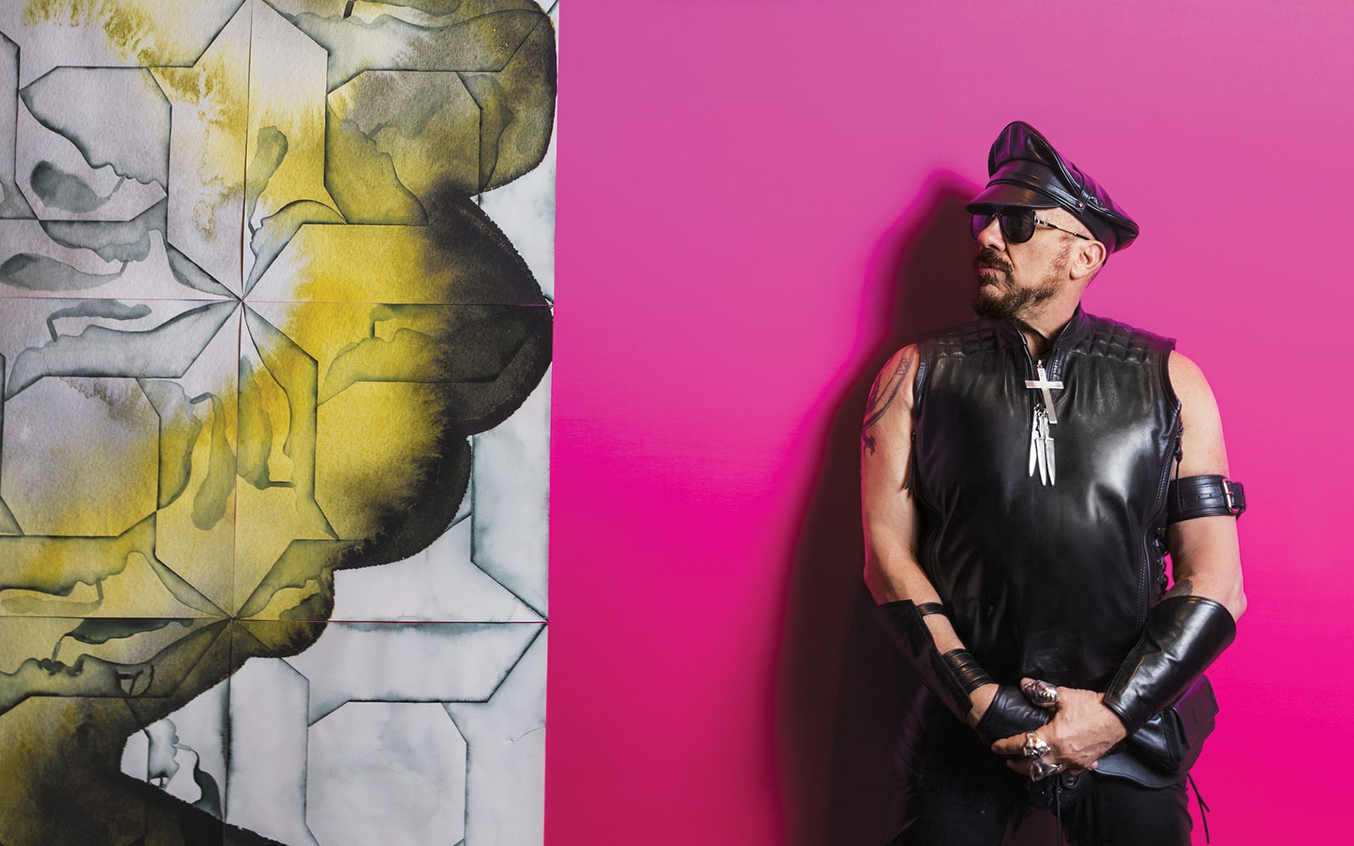 A person in black leather attire and sunglasses stands against a pink wall next to a yellow and gray abstract painting.