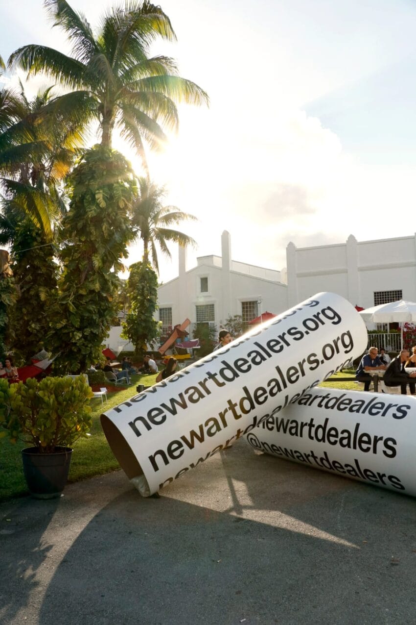 Large cylindrical sign displaying newartdealers.org at an outdoor event with palm trees and a white building in the background.