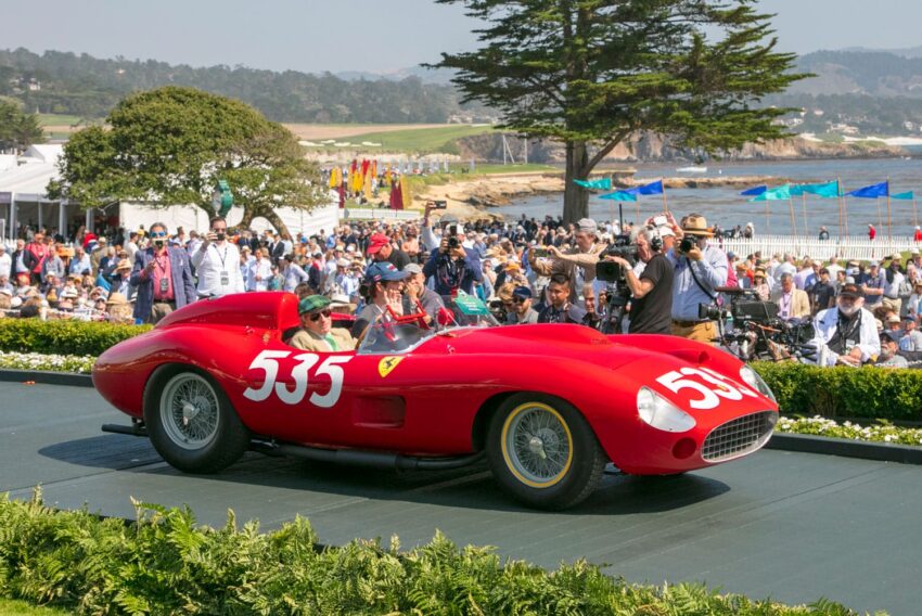 Red vintage sports car with number 535, driven by a person in a cap, surrounded by a crowd at an outdoor event.