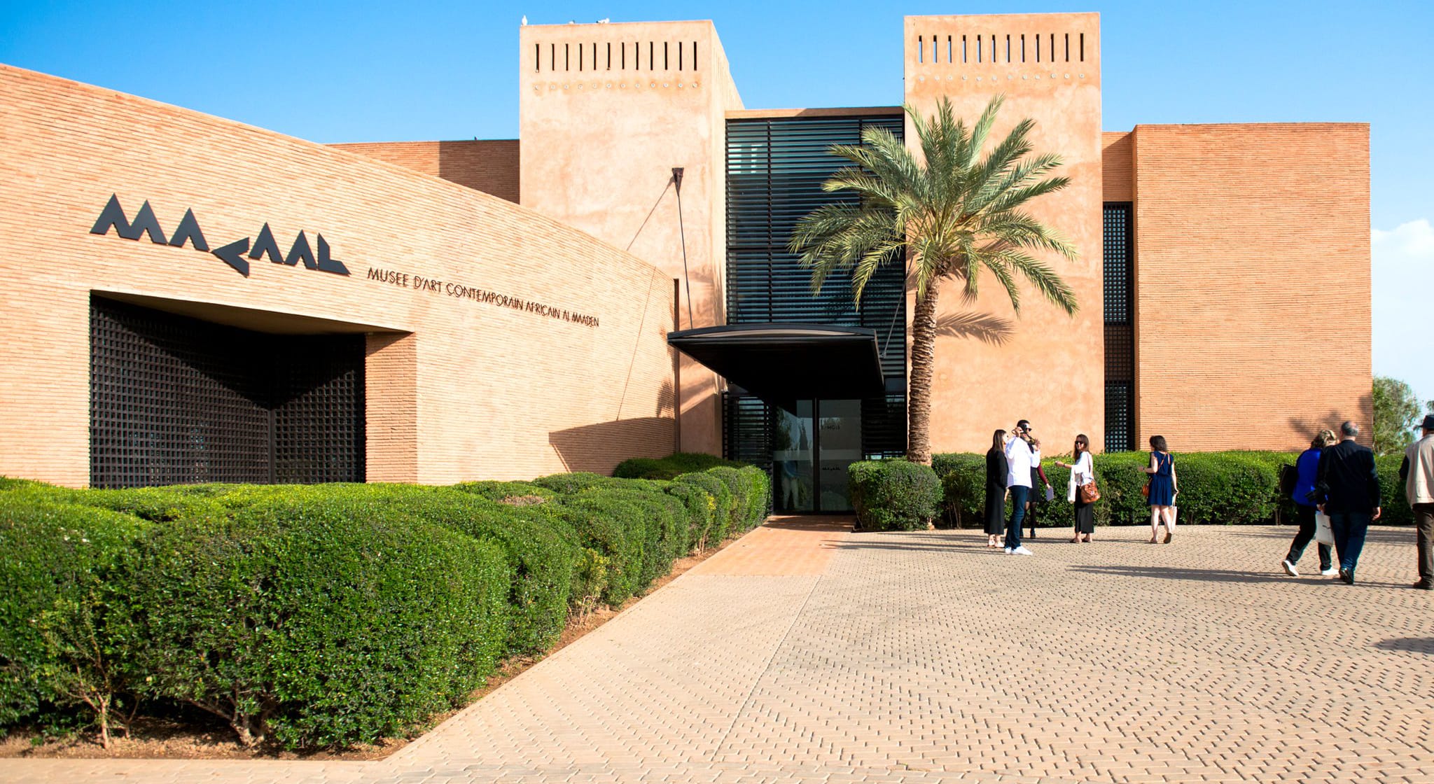 Exterior view of a modern art museum with brick walls, palm tree, and people walking on a pathway under a clear blue sky.
