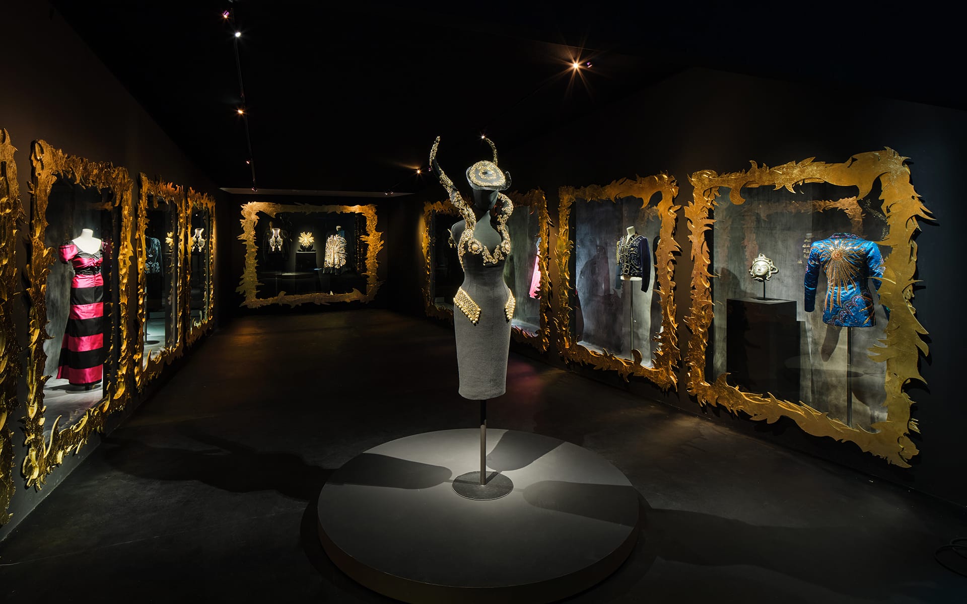 Fashion exhibit with ornate frames displaying various outfits in a dimly lit room with a central mannequin wearing a hat.