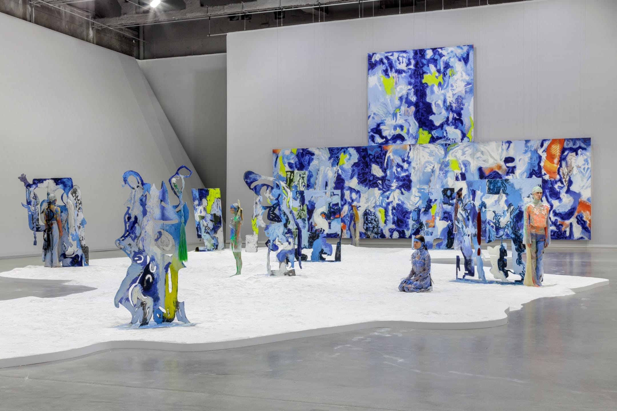 Contemporary art installation with colorful abstract sculptures and large blue and white canvases in a spacious gallery.