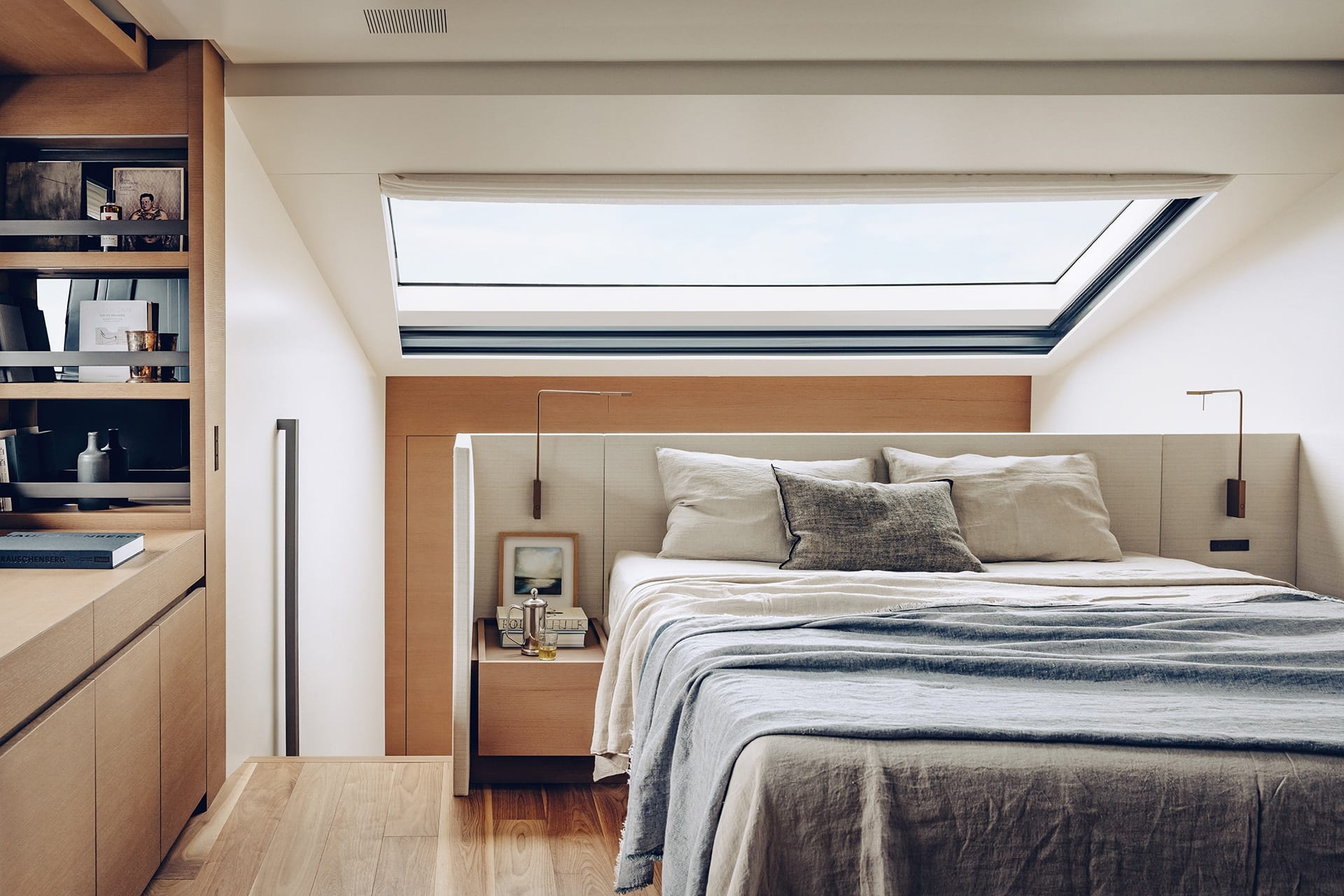 Modern bedroom with skylight, wooden accents, cozy bed, pillows, blankets, books on shelf, natural light filling the room.