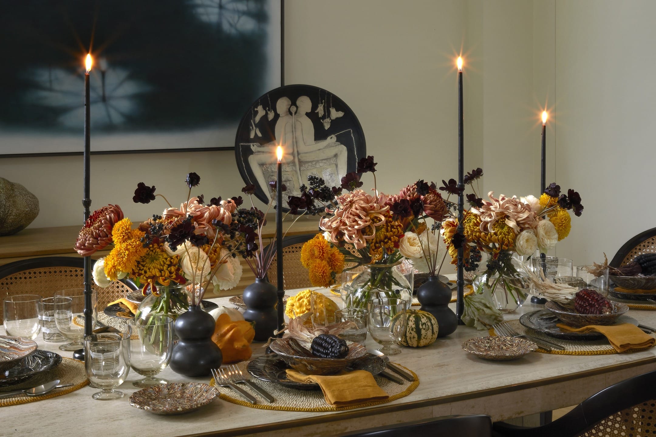Elegant dining table set with autumnal floral arrangements, dark candles, fine china, and warm-toned napkins.