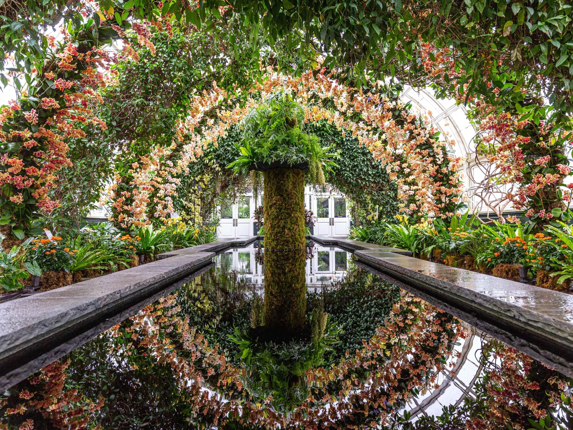 Ornate greenhouse interior with colorful flower arches, lush greenery, and reflective water feature creating a serene ambiance.