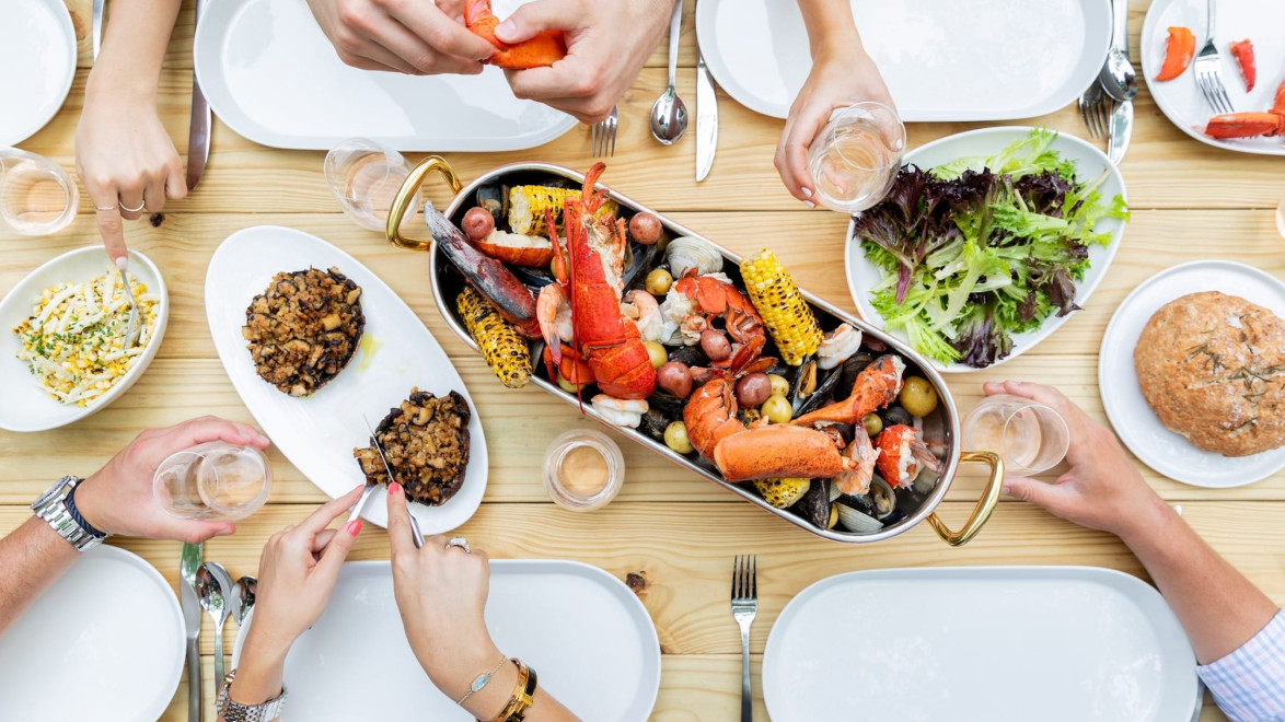 People enjoying a seafood feast at a dining table with lobster, corn, salad, and various dishes.