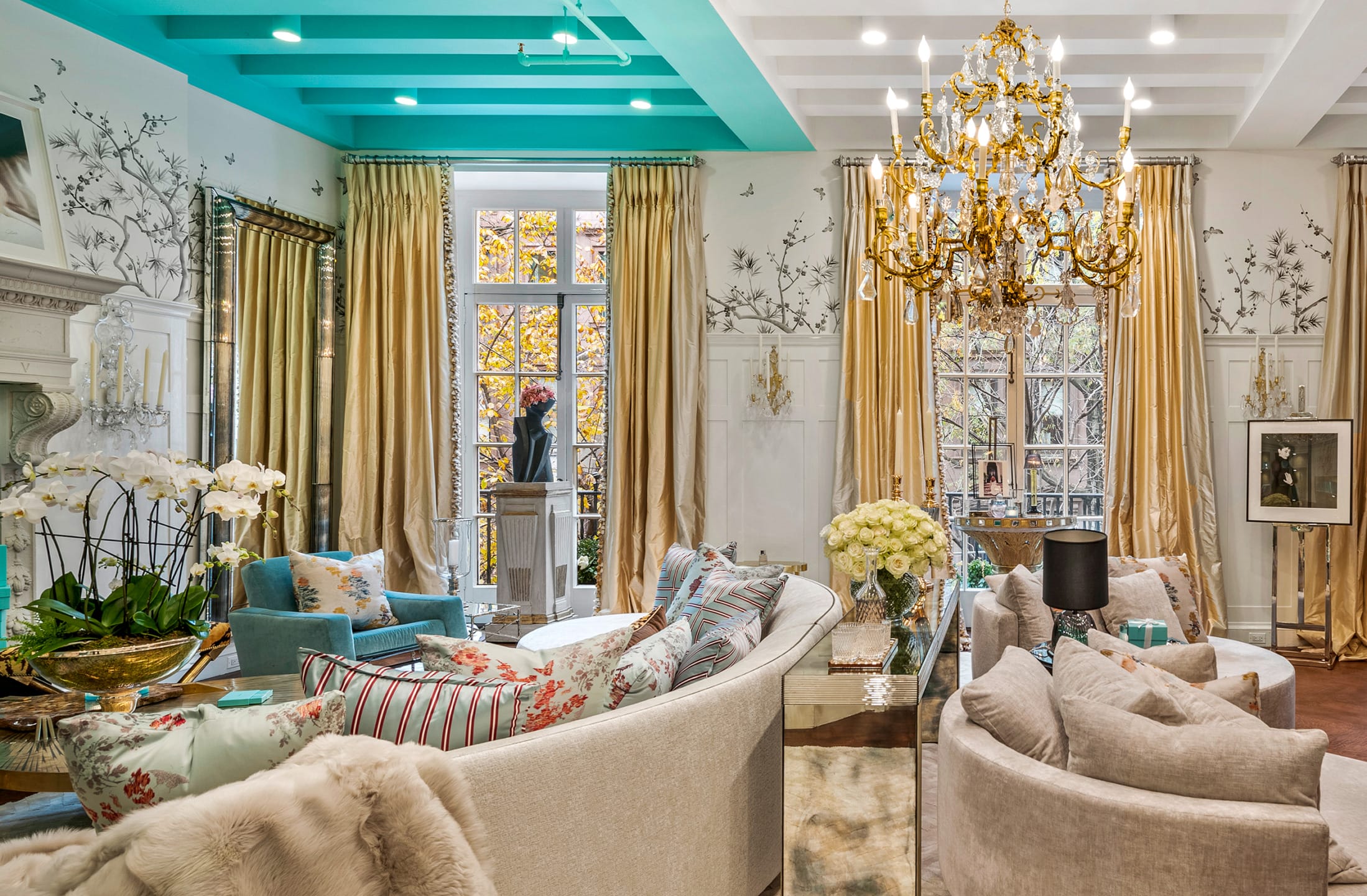 Elegant living room with floral sofas, gold chandelier, turquoise ceiling, and large windows with gold curtains.